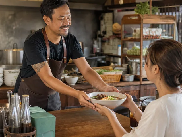 Cook serving a boat noodle bowl to a diner in a Thai kitchen